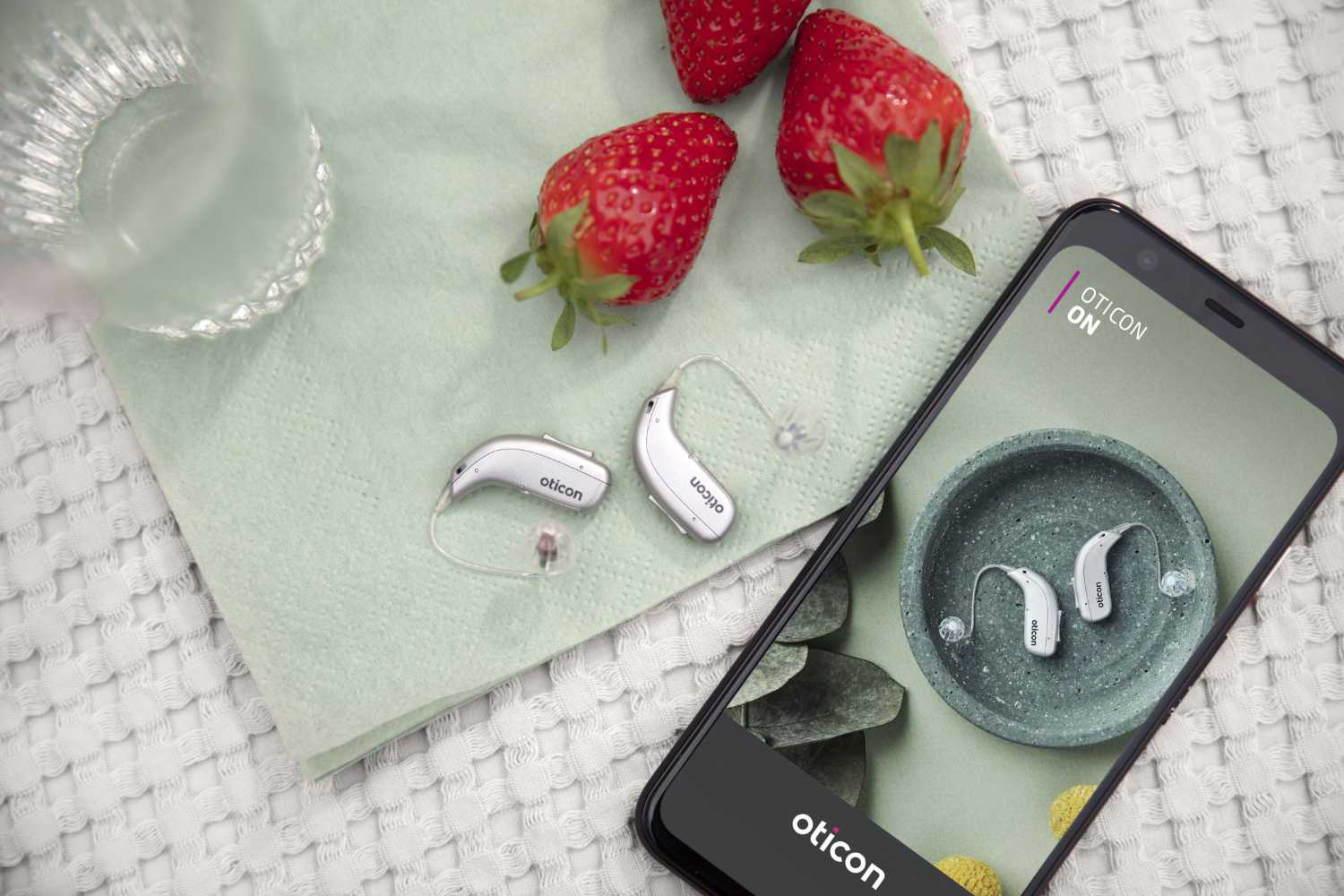 Hörgerätepaar der Firma Oticon, die neben einem Smartphone mit geöffneter Oticon-App liegen