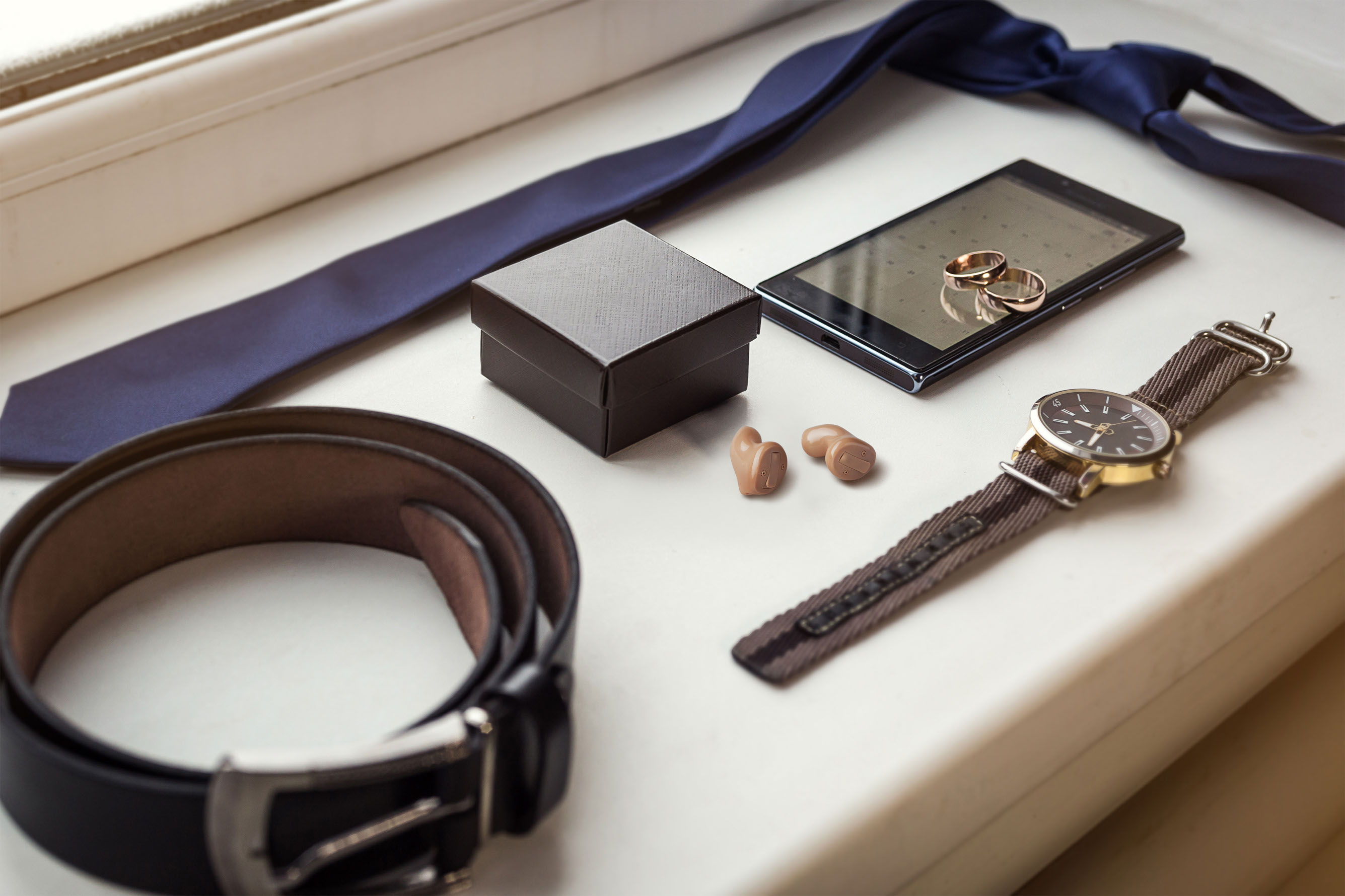 zwei winzige, teilweise transparente Hörgeräte zwischen männlichen Luxusartikeln wie Gürtel, Krawatte, Armbanduhr und Ringen